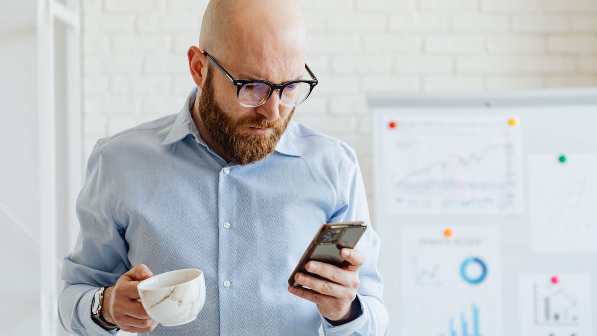 A man in glasses checking his phone while holding a coffee cup indoors