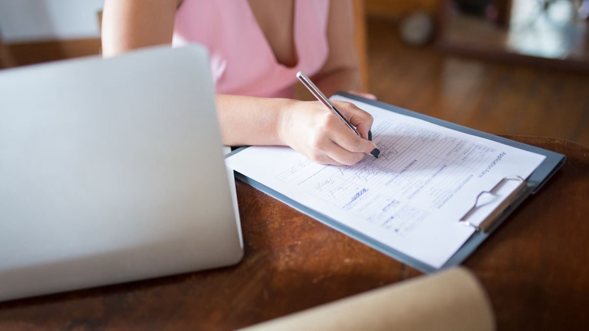 A woman writing on a document on a clipboard with a laptop beside her