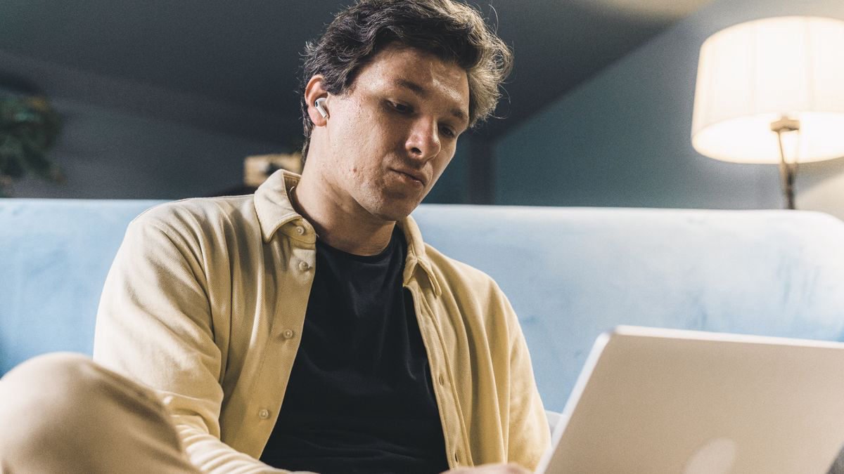 Young man sitting indoors working on a laptop with a thoughtful expression
