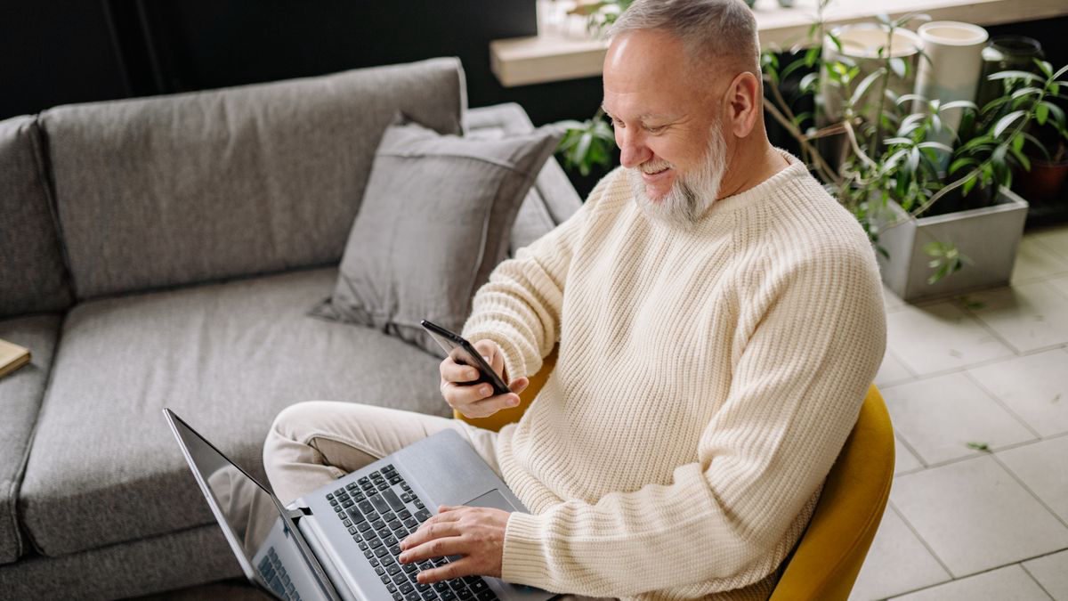 Smiling older man at home using a phone and a laptop