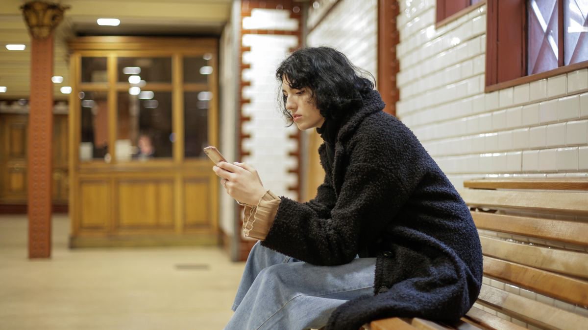 A young woman in a black coat sitting on a bench looking at her phone