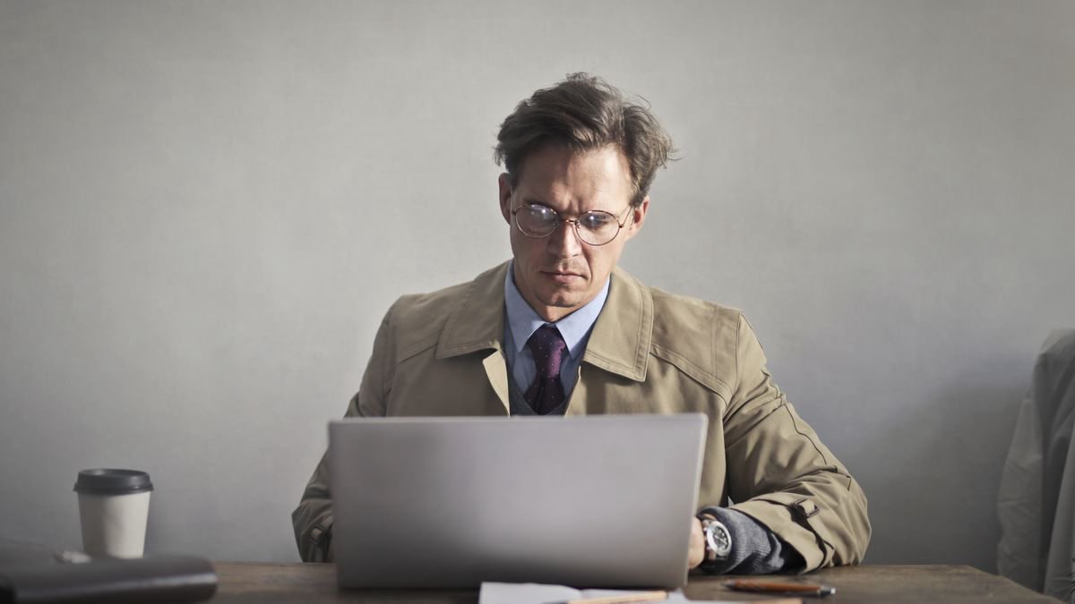 A man in glasses working on a laptop at a wooden desk