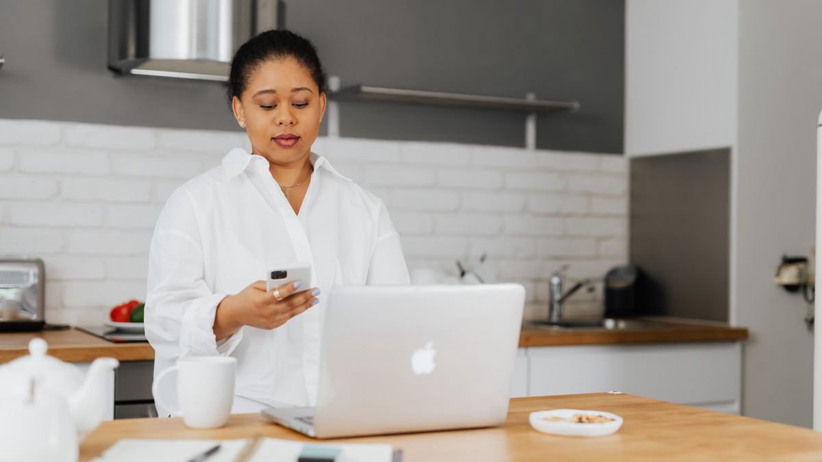 A woman using her smartphone in a modern kitchen next to an open laptop