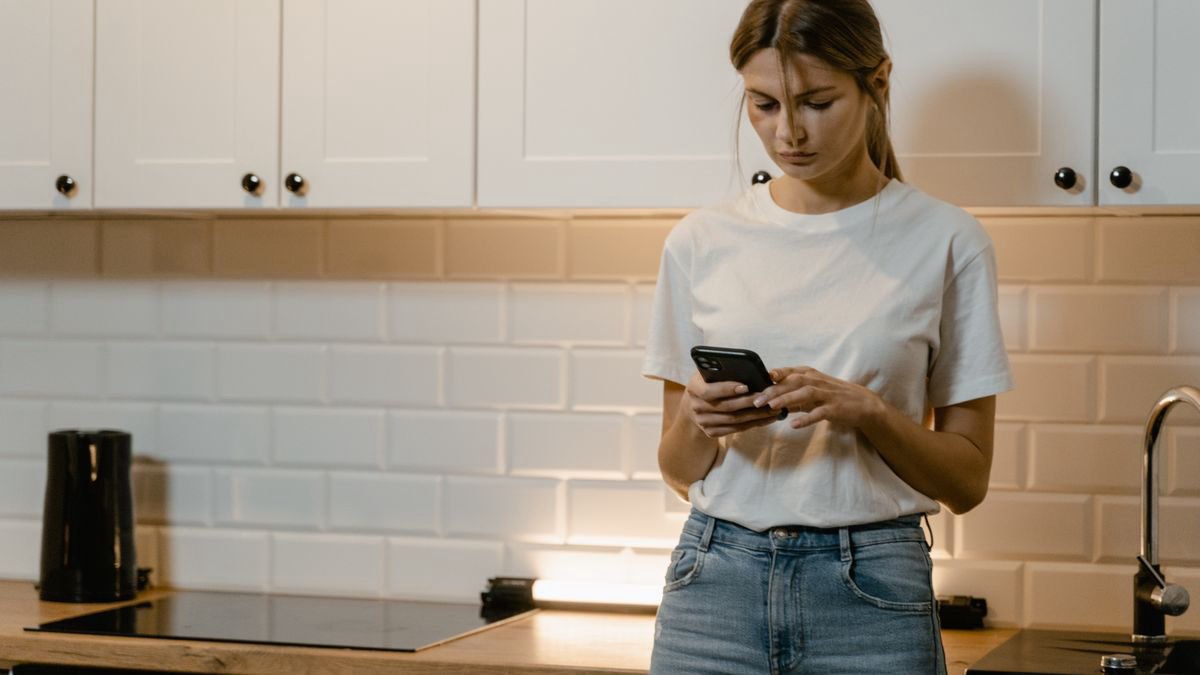 A young woman in a kitchen looking thoughtfully at her smartphone