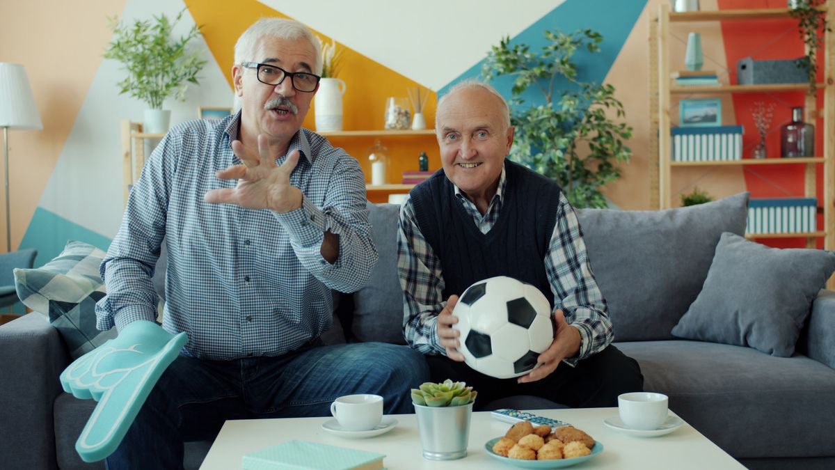 Two older men watching a soccer game at home with snacks