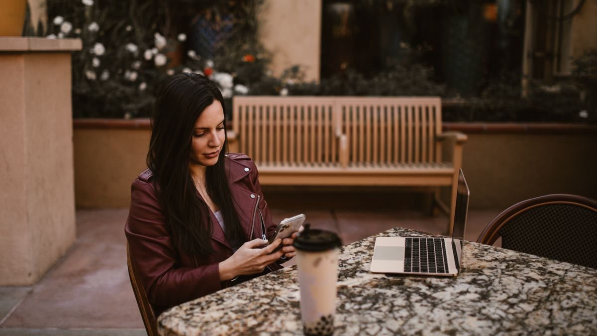 Woman checking her smartphone at an outdoor cafe with a laptop and coffee