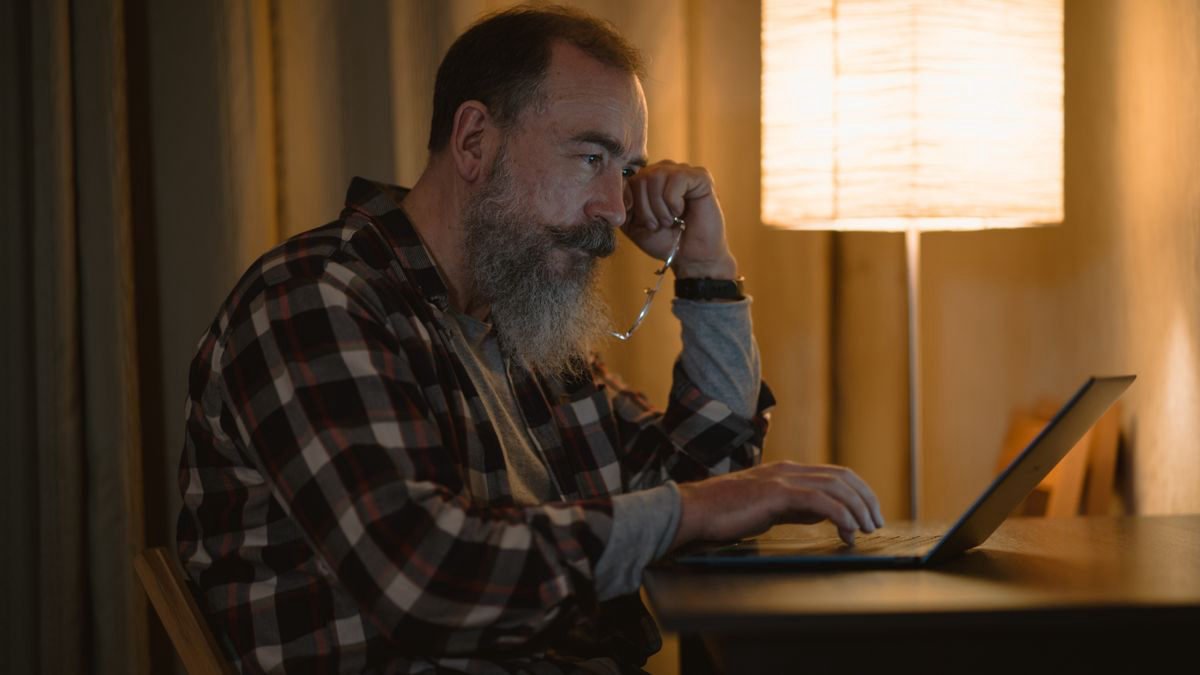 Older man with a beard working on a laptop under warm lamp light