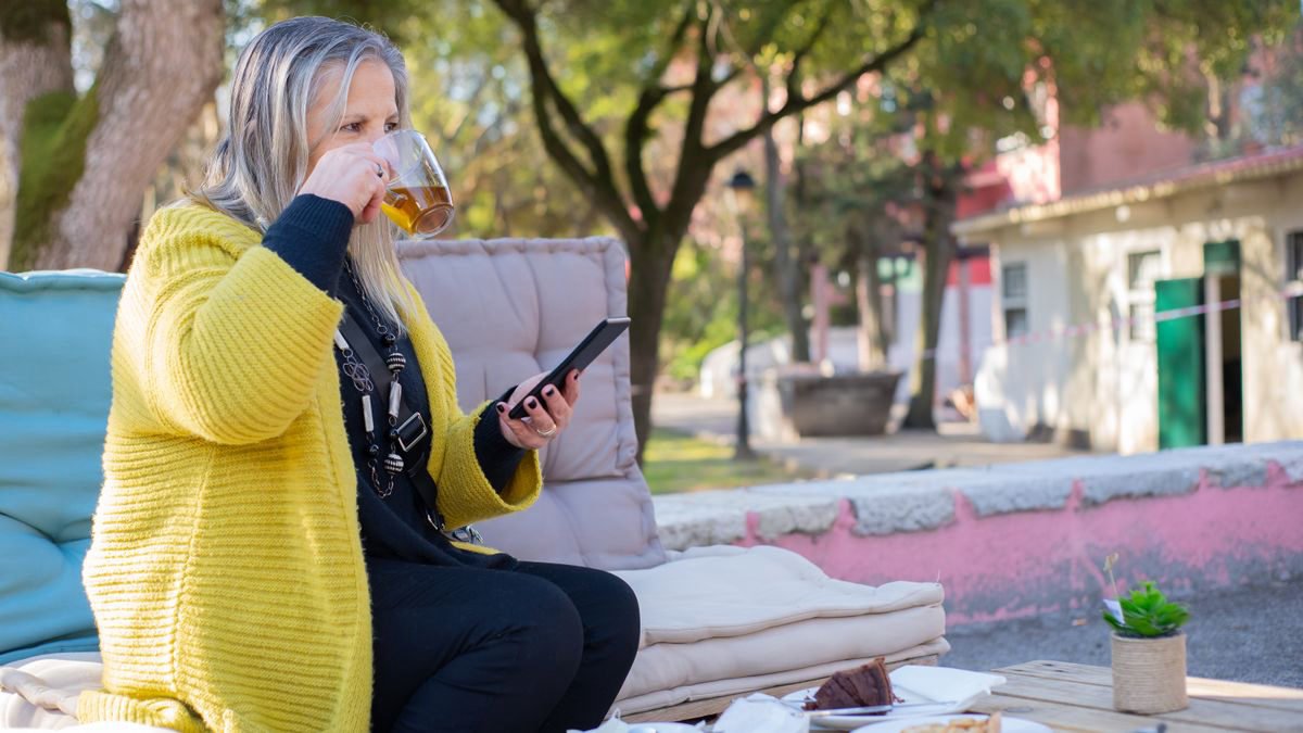 Senior woman in a yellow sweater drinking tea and using a smartphone outdoors
