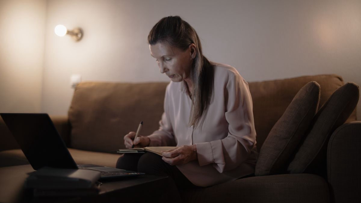 Senior woman on a couch writing notes while using a laptop