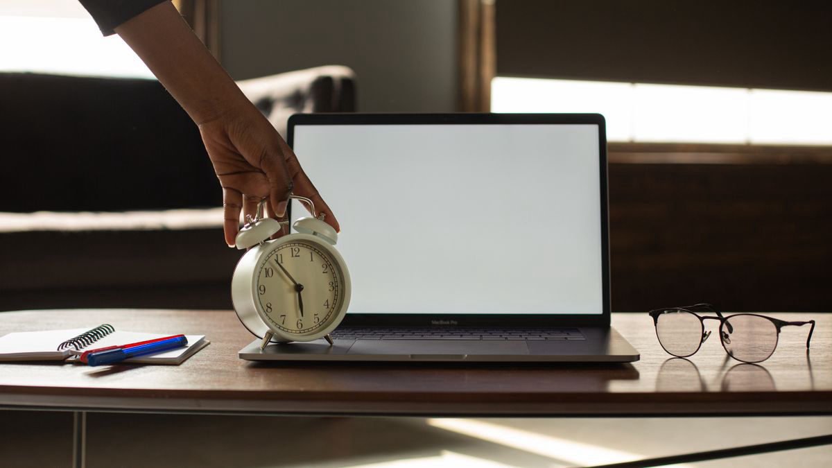 A person reaching for an alarm clock next to an open laptop