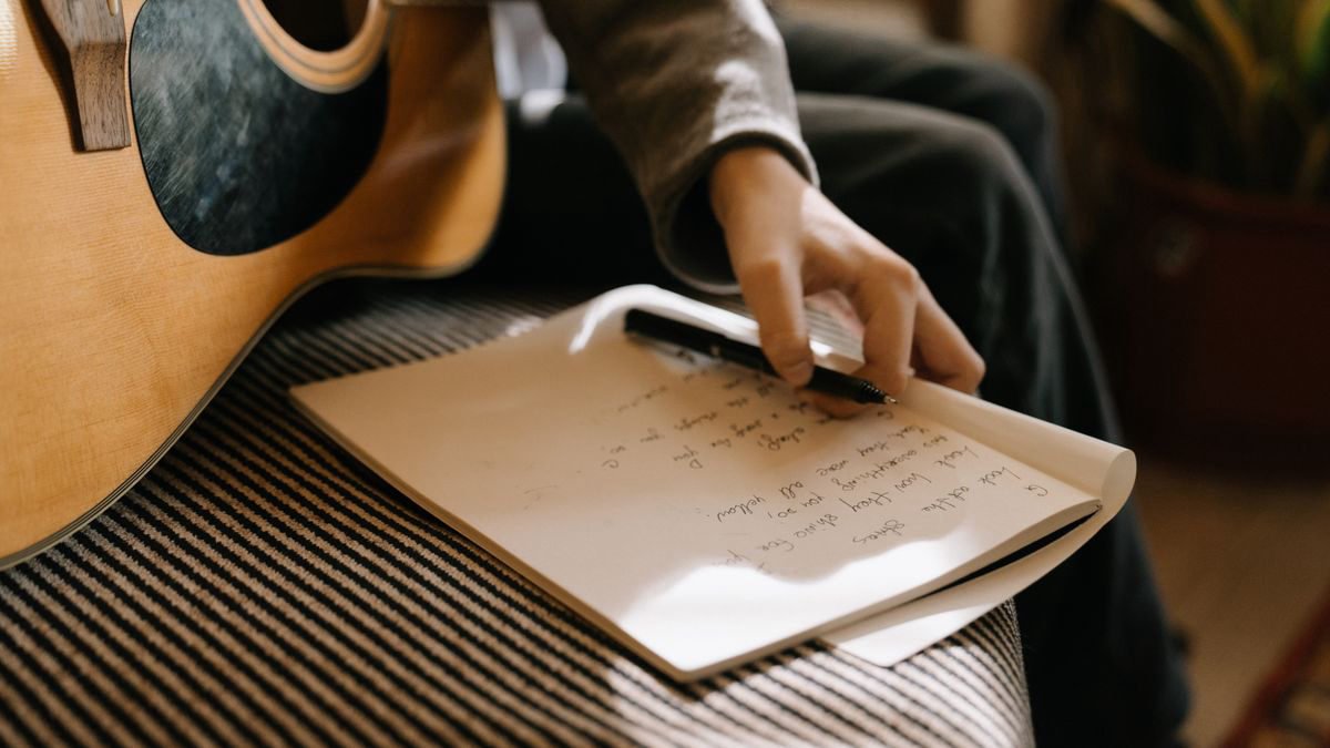 Person writing song lyrics in a notebook with an acoustic guitar