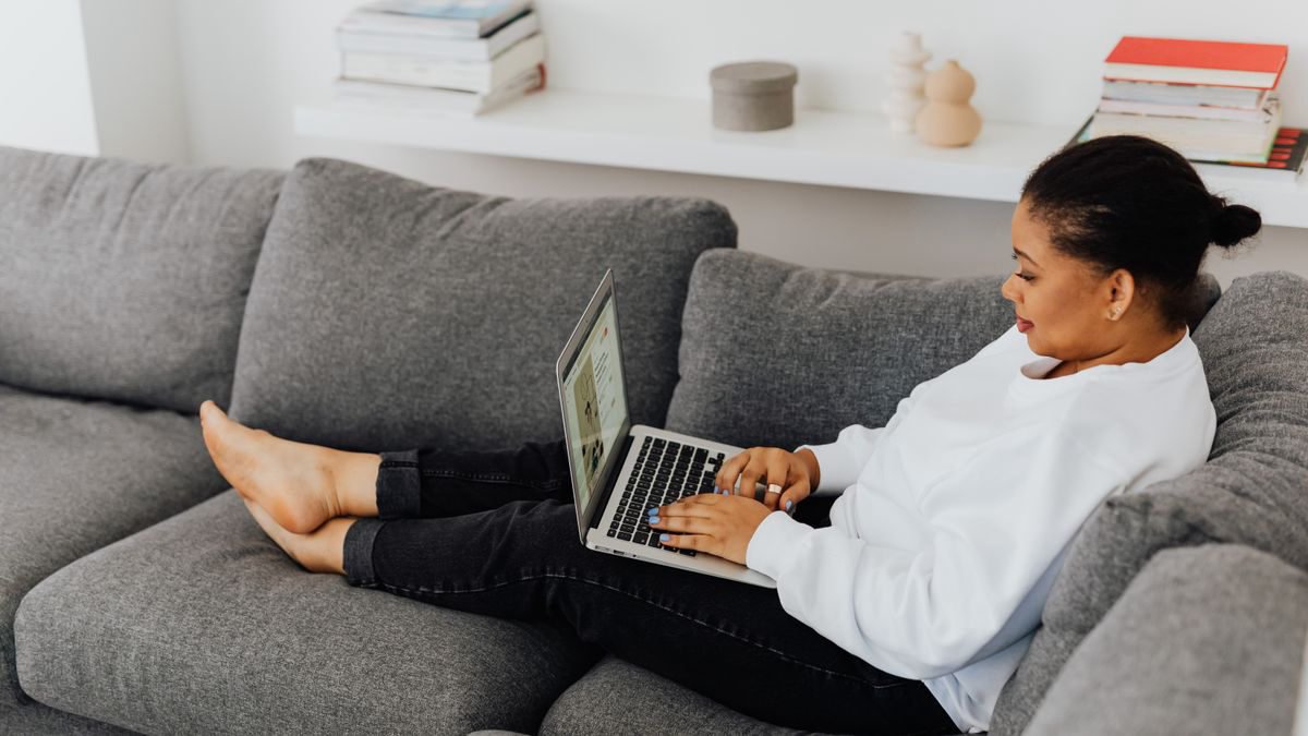 Woman in a white sweater using a laptop while lying on a sofa